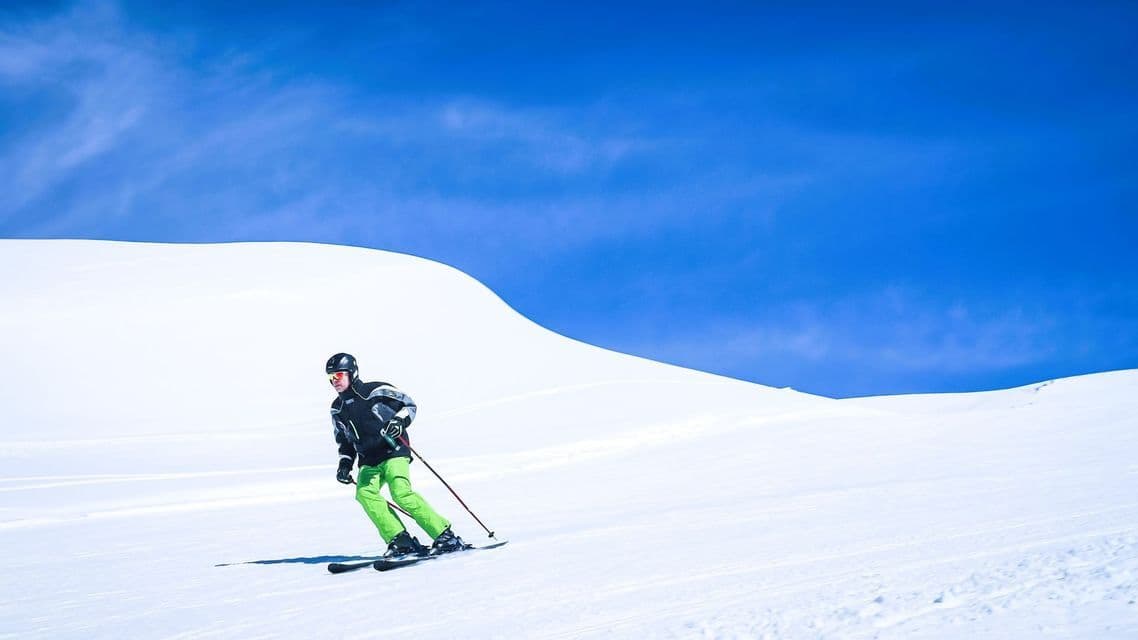 Ein Skifahrer mit Helm und leuchtend grüner Hose gleitet unter klarem blauem Himmel einen schneebedeckten Berg hinunter.