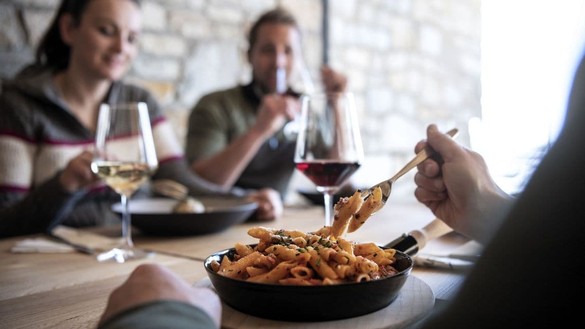 Nahaufnahme von Penne-Pasta, die aus einer Schüssel gegessen wird, während eine WeRoad-Gruppenreise im Hintergrund speist und Wein trinkt.