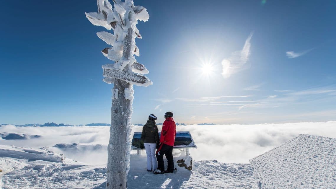 Zwei Personen in Skiausrüstung stehen auf einem verschneiten Berggipfel und blicken unter strahlender Sonne auf ein Wolkenmeer.