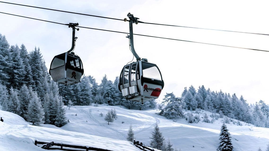 Zwei Skilift-Gondeln schweben an einem Seil über einer verschneiten Berglandschaft mit schneebedeckten Tannen.