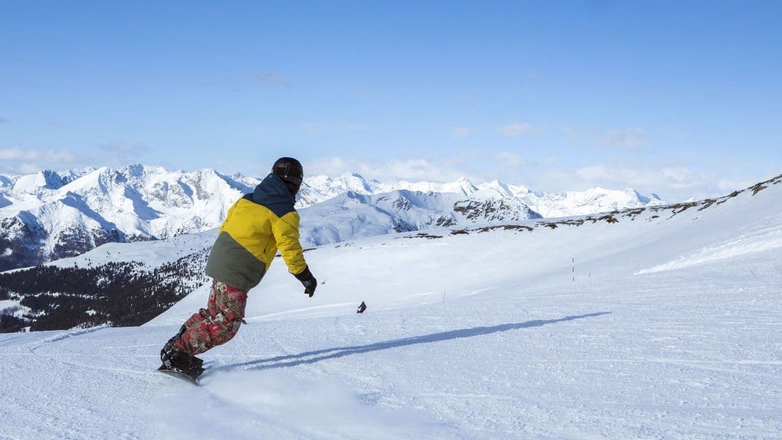 Eine Person in einer gelben Jacke snowboardet einen verschneiten Hang hinunter, mit einer weiten Kette schneebedeckter Berge im Hintergrund unter einem klaren Himmel.