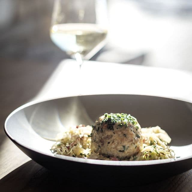 Nahaufnahme eines großen, mit Kräutern garnierten Knödels in einer dunklen Schale, mit einem Glas Weißwein im Hintergrund.