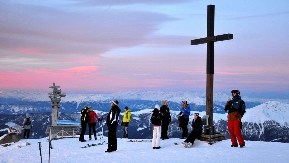 WeRoad Gruppenreise auf einem verschneiten Berggipfel bei Sonnenaufgang, nahe einem großen Kreuz mit schneebedeckten Gipfeln im Hintergrund.