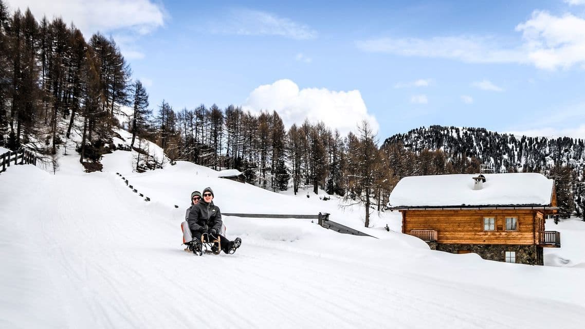 Zwei Personen in Winterkleidung rodeln einen verschneiten Hügel hinunter, vorbei an einer Holzhütte und einem Kiefernwald.