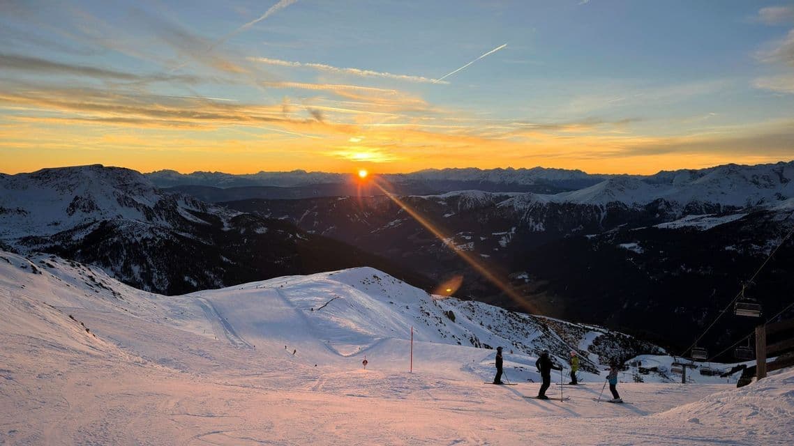 Eine WeRoad-Gruppe von Skifahrern pausiert auf einem verschneiten Berghang und genießt den Sonnenuntergang hinter fernen Gipfeln.
