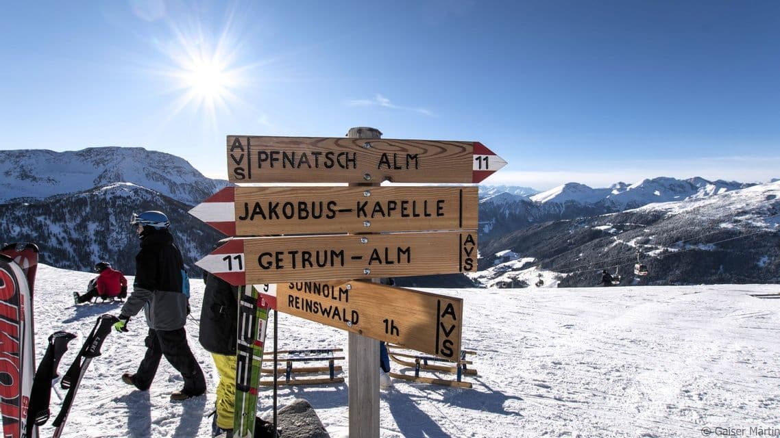 Ein hölzerner Wegweiser an einem verschneiten Berghang mit Skifahrern und sichtbaren fernen Gipfeln unter strahlender Sonne und klarem blauem Himmel.