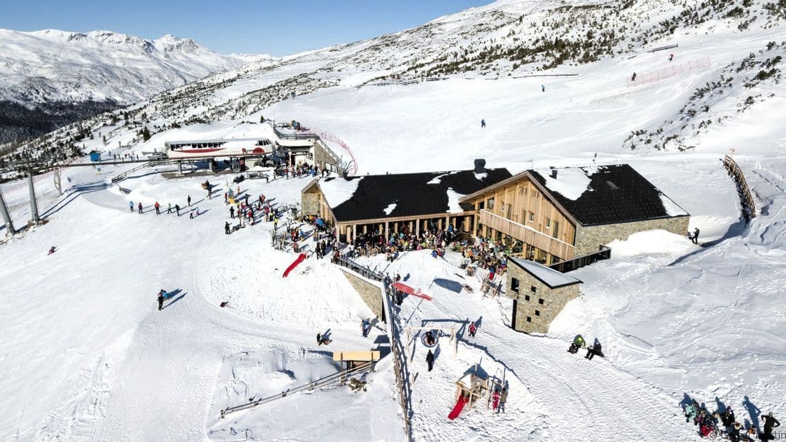 Luftaufnahme einer belebten Skihütten-Lodge mit Menschen auf der Terrasse und Skifahrern auf den umliegenden verschneiten Hängen unter klarem Himmel.