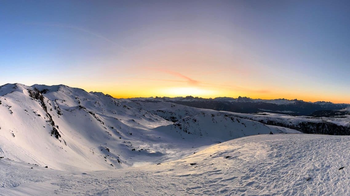 Ein Panoramablick auf eine schneebedeckte Bergkette bei einem goldenen Sonnenuntergang unter einem klaren, farbverlaufenden Himmel.