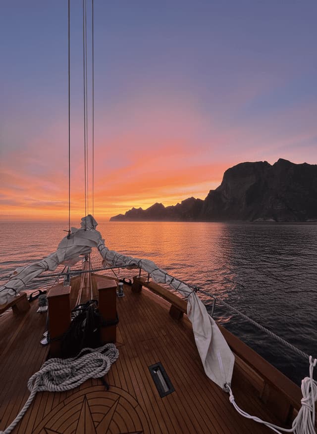 Der Blick vom Holzdeck eines Segelboots auf eine bergige Küste bei einem farbenfrohen Sonnenuntergang.
