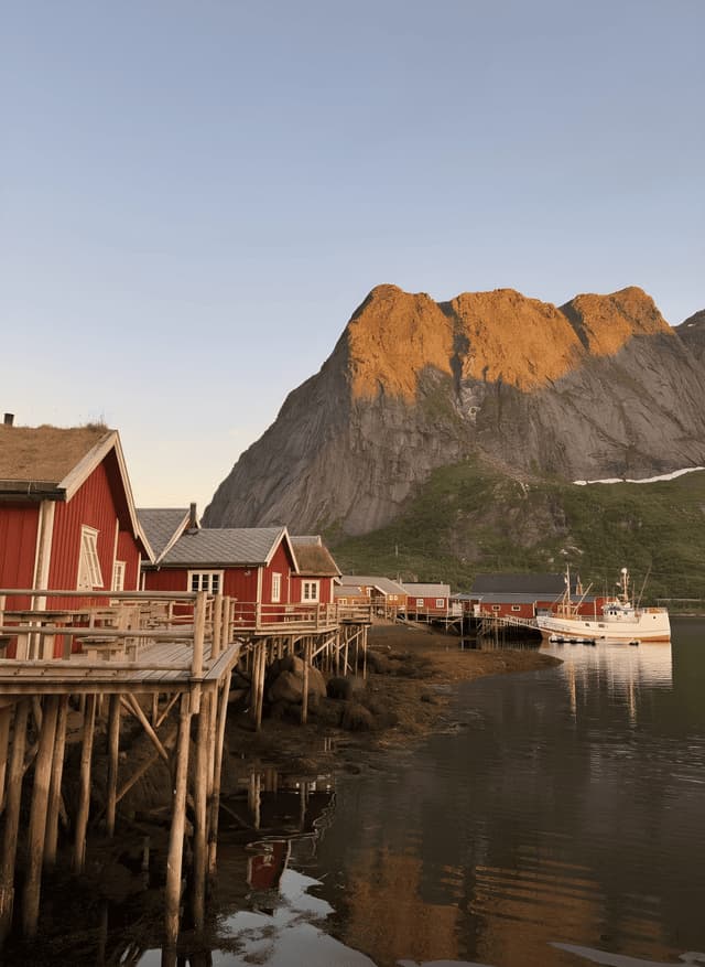 Capanne di legno rosse su palafitte costeggiano una baia tranquilla ai piedi di una grande montagna illuminata dalla luce dorata del sole.