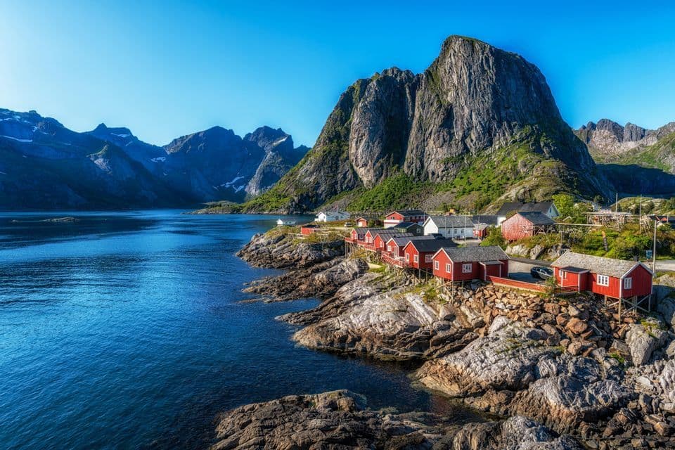 Un villaggio marittimo con case di legno rosse costruite su una costa rocciosa accanto a un fiordo, con ripide montagne sullo sfondo sotto un cielo sereno.