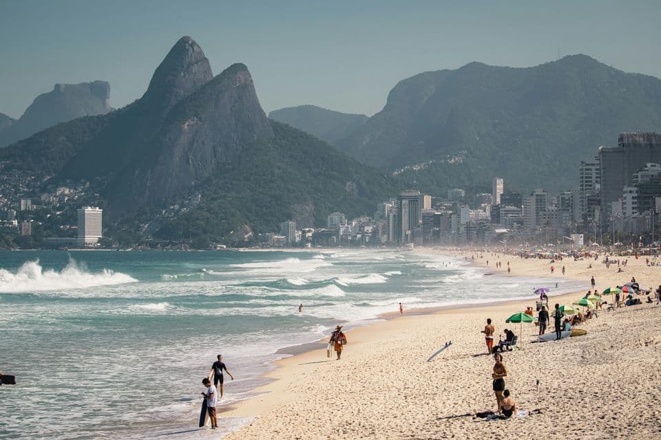 Una vista amplia de una playa urbana concurrida con gente en la arena, con el horizonte de una ciudad y grandes montañas cubiertas de árboles al fondo.