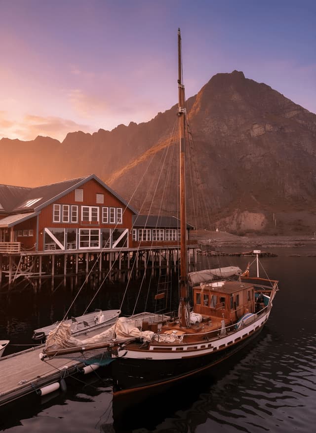 Ein hölzernes Segelboot liegt an einem Pier neben einem roten Gebäude auf Stelzen, mit einem großen Berg im Hintergrund bei Sonnenuntergang.