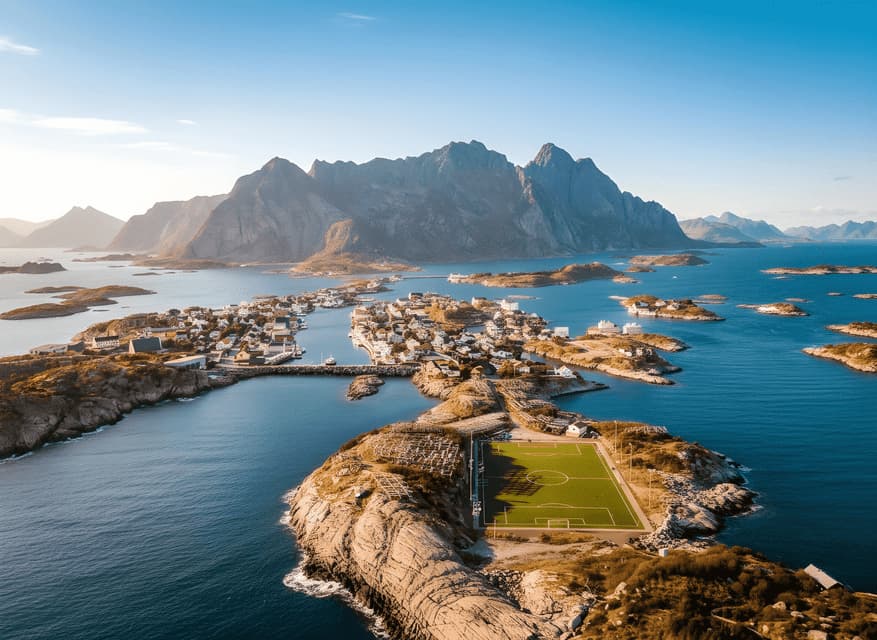 Eine Luftaufnahme eines Küstendorfes und eines Fußballfeldes, die auf felsigen Inseln liegen, mit großen Bergen, die im Hintergrund aus dem Meer ragen.