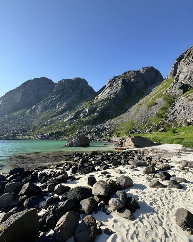 Una spiaggia sabbiosa e rocciosa costeggia acque calme e turchesi ai piedi di grandi montagne verdi sotto un cielo azzurro e limpido.