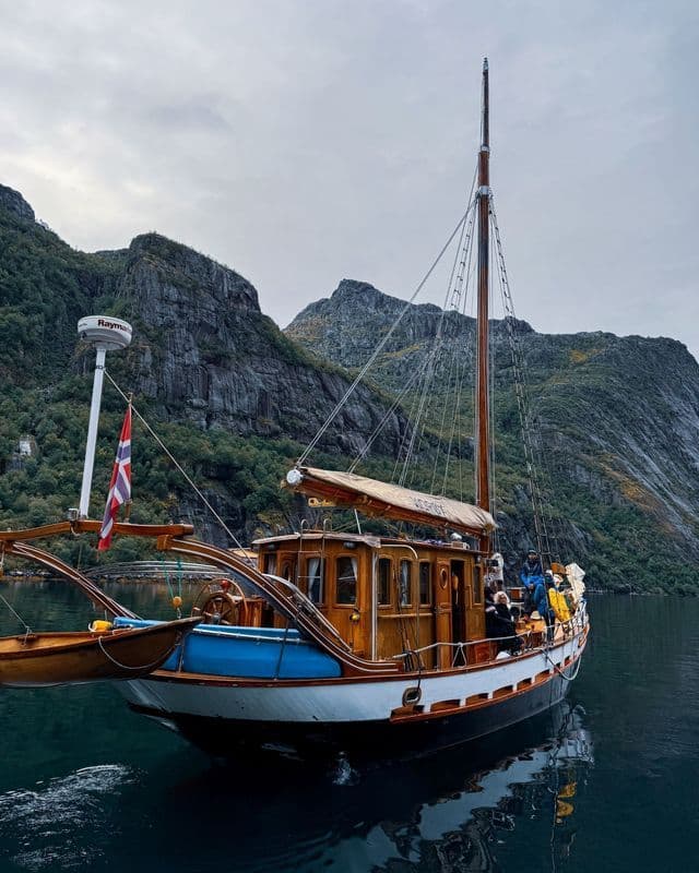 Eine WeRoad Gruppenreise auf einem Holzsegelboot fährt entlang eines Fjords mit steilen, felsigen Bergen im Hintergrund.