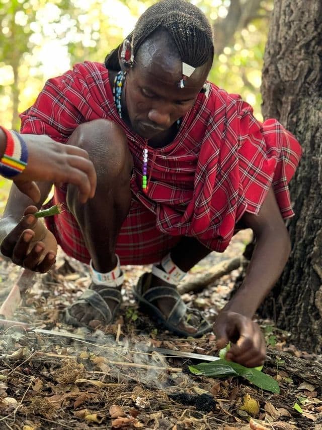 Un uomo in abiti tradizionali Maasai si accovaccia in una foresta, accudendo un piccolo fuoco fumante a terra con delle foglie.
