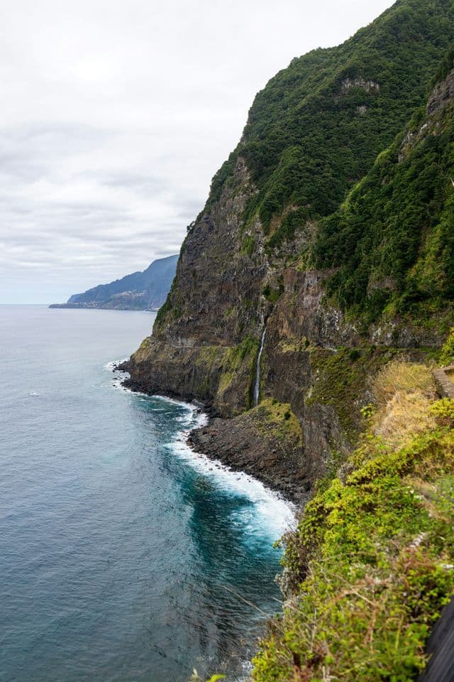 Una sottile cascata scende da una ripida scogliera ricoperta di verde nell'oceano agitato sottostante, con una costa frastagliata in lontananza.