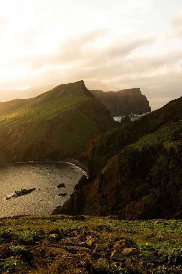 Acantilados escarpados y cubiertos de verde se adentran en el océano, rodeando una bahía tranquila bajo la cálida luz del sol.