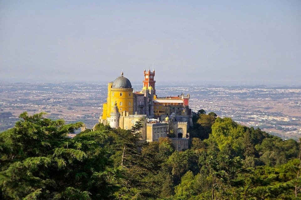 Un castello variopinto con torri gialle e rosse si erge su una collina boscosa, dominando un vasto paesaggio.