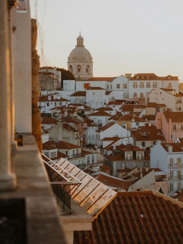 Une vue plongeante d'une ville aux bâtiments blancs et aux toits de tuiles rouges, avec un grand dôme de pierre visible au loin au coucher du soleil.