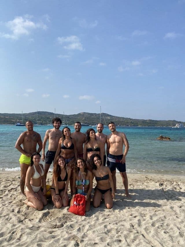 Un viaggio di gruppo WeRoad con uomini e donne in costume da bagno in posa per una foto su una spiaggia sabbiosa, con acqua turchese e barche a vela sullo sfondo.