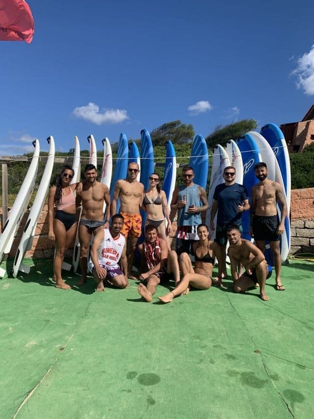Eine WeRoad-Gruppe in Badekleidung posiert vor einer Reihe von Surfbrettern unter strahlend blauem Himmel für ein Foto.