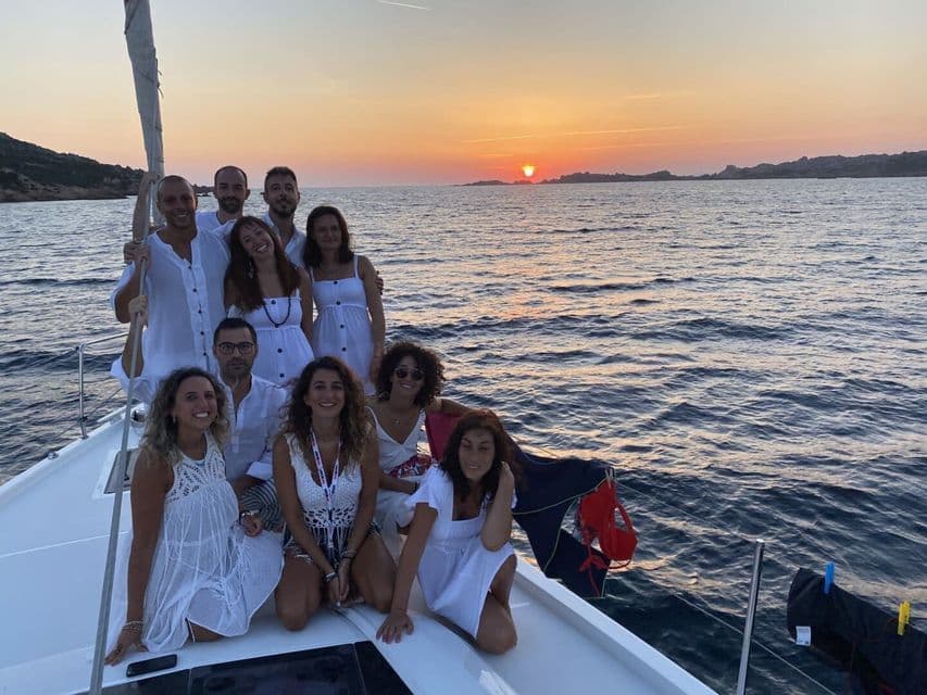 Eine WeRoad Gruppenreise, alle in Weiß gekleidet, posieren zusammen an Deck eines Segelboots bei Sonnenuntergang.