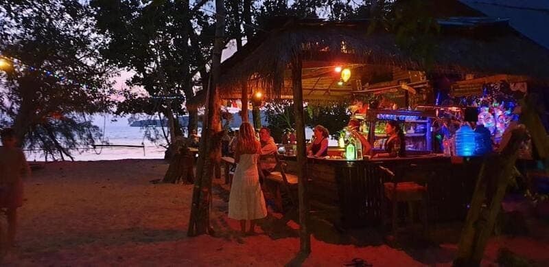 Un voyage en groupe WeRoad dans un bar de plage illuminé sur le sable au crépuscule, avec la mer en toile de fond.