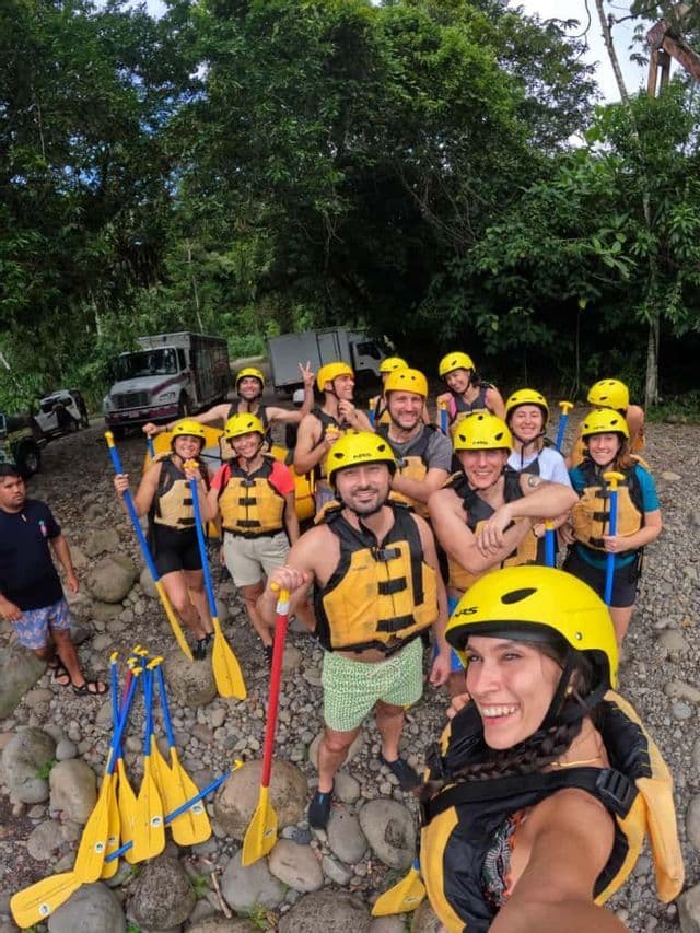 Un gruppo WeRoad con caschi gialli e giubbotti di salvataggio posa per un selfie su una riva rocciosa del fiume prima di fare rafting.