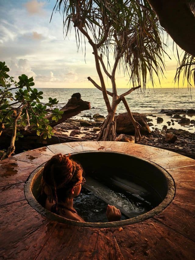Une personne aux dreadlocks se prélasse dans un bain en bois extérieur sur une côte rocheuse, contemplant le coucher de soleil sur la mer.