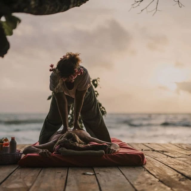 Une personne avec des fleurs dans les cheveux donne un massage du dos à quelqu'un allongé sur un tapis sur une terrasse en bois en bord de mer au coucher du soleil.