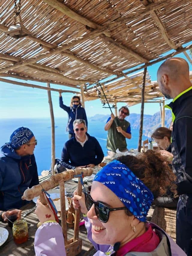 Un viaggio di gruppo WeRoad si riunisce attorno a un tavolo di legno sotto un tetto rustico di bambù, con una vista panoramica sul mare e sulla costa.