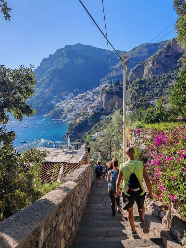 Eine WeRoad-Gruppenreise, die eine Steintreppe hinabsteigt und eine Klippenstadt sowie das blaue Meer überblickt.