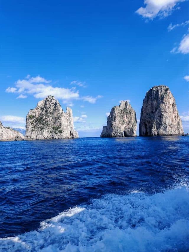 Große Felsformationen erheben sich aus dem tiefblauen Meer unter einem sonnigen Himmel, mit der weißen Bugwelle eines Bootes im Vordergrund.
