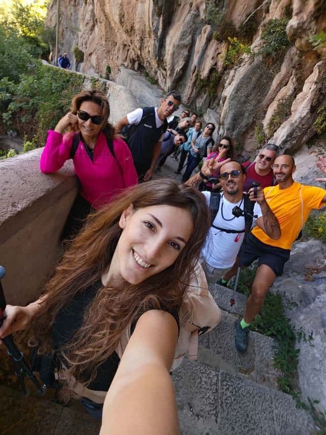 Una donna sorridente si scatta un selfie durante un viaggio di gruppo WeRoad, posando su una ripida scalinata in pietra incastonata in una scogliera rocciosa.