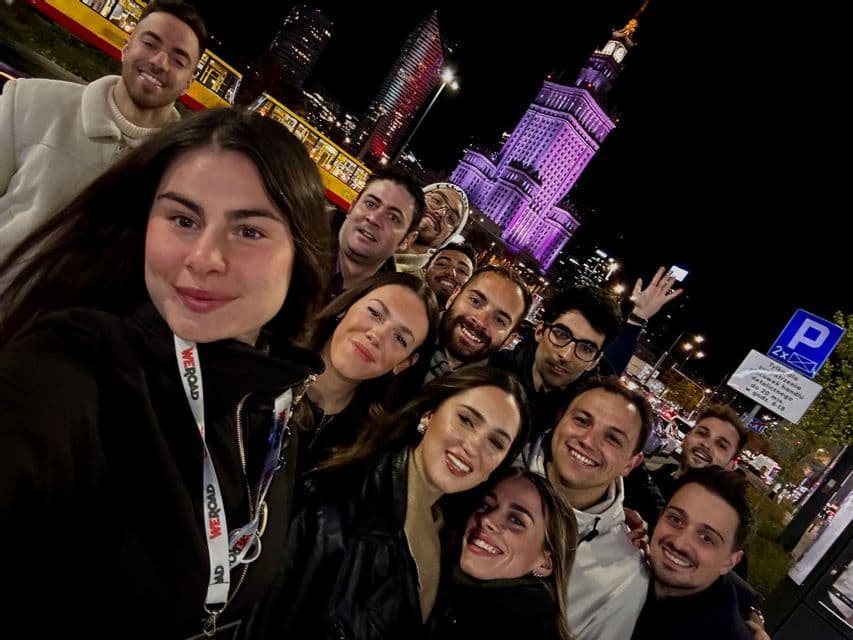 Un gruppo WeRoad si scatta un selfie sorridendo di notte in una via di città, con un edificio illuminato e un tram sullo sfondo.