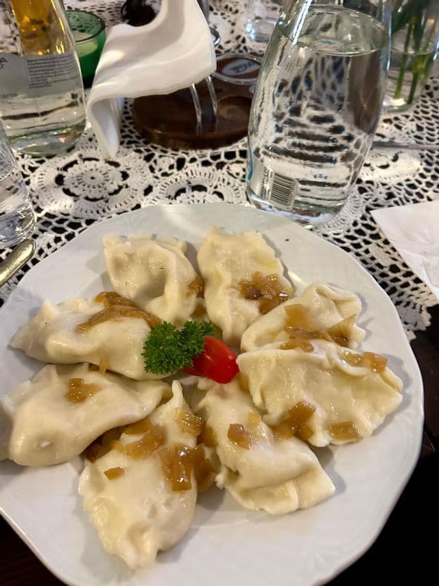 Une assiette blanche de pierogi garnis d'oignons caramélisés, décorée de persil, posée sur une nappe en dentelle.