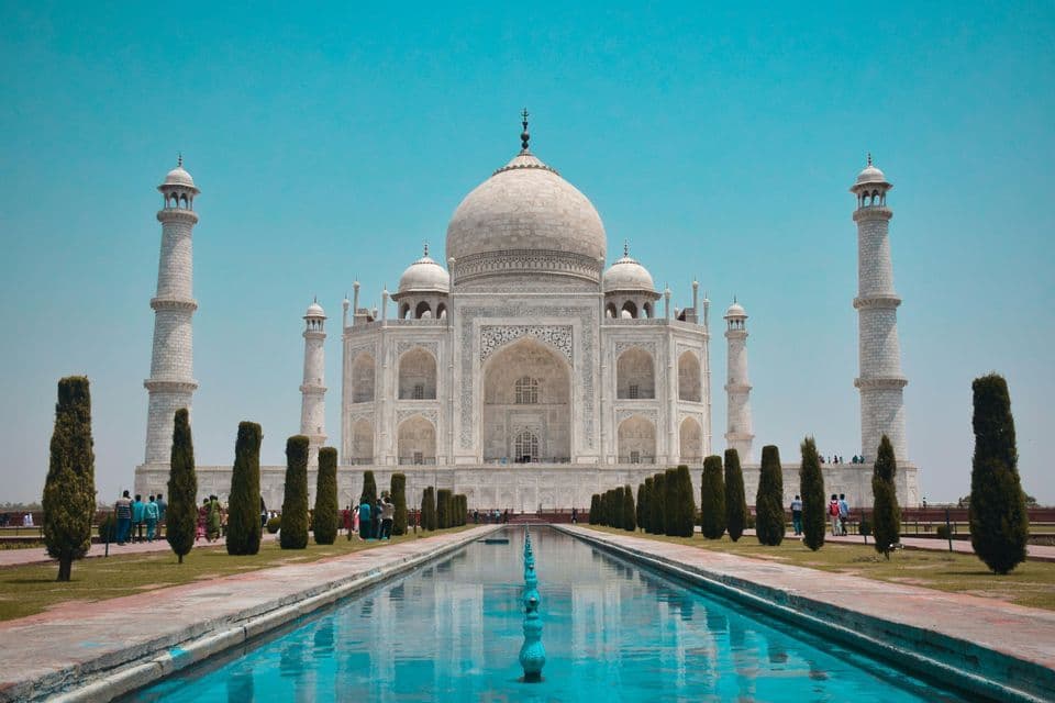 Il Taj Mahal, un mausoleo di marmo bianco, si riflette nella lunga vasca dei suoi giardini sotto un cielo azzurro e limpido.