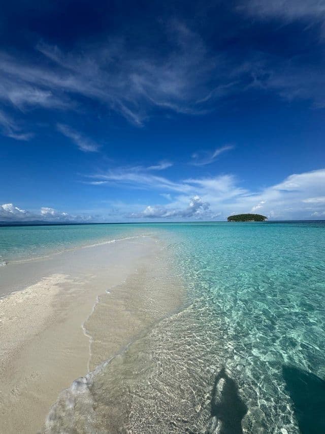 Una lingua di sabbia bianca incontra acque turchesi e cristalline sotto un cielo azzurro, con l'ombra di una persona in primo piano e una piccola isola all'orizzonte.