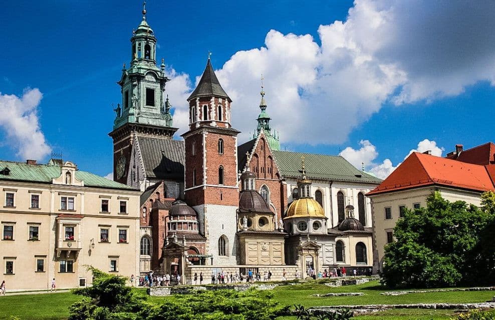 A historic cathedral complex with brick towers, green spires, and a golden dome sits on a grassy hill under a blue sky with clouds.