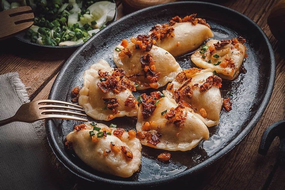 A close-up of pierogi dumplings garnished with fried onions and chives, served on a black plate with a fork.