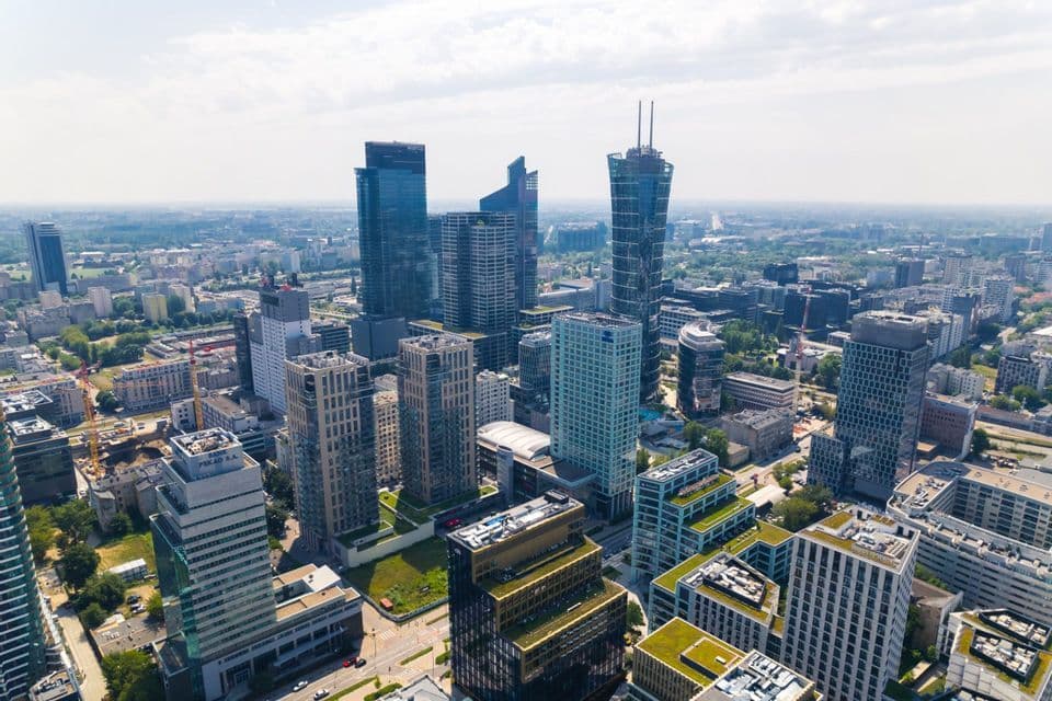 An aerial view of a dense modern cityscape with glass skyscrapers and green-roofed buildings under a bright, partly cloudy sky.