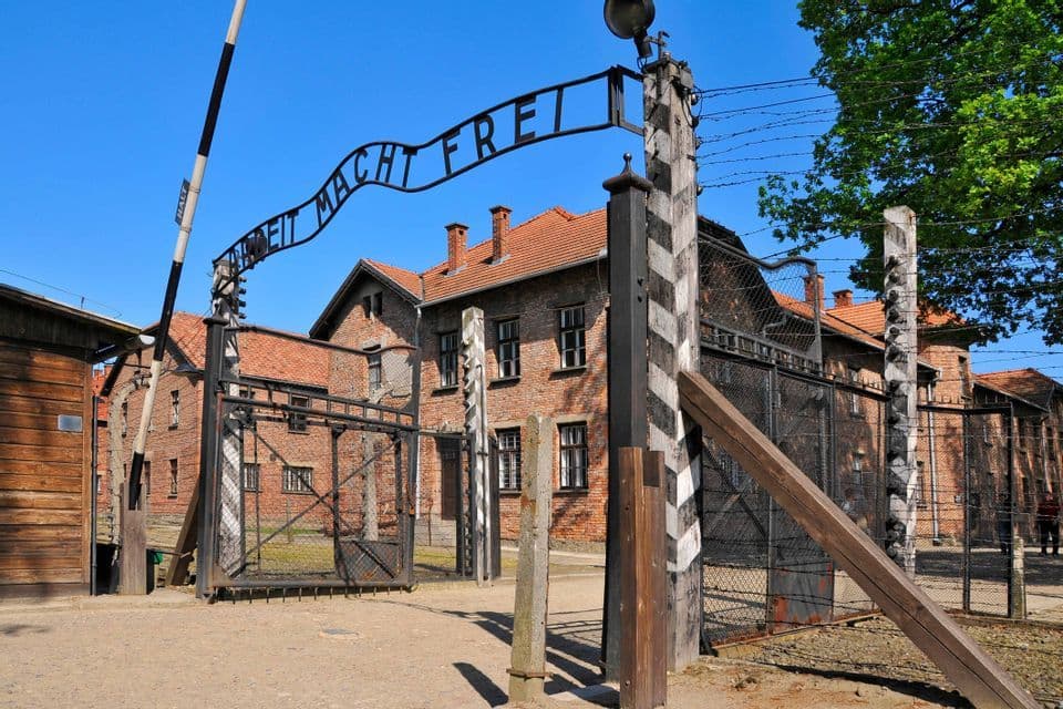 L'insegna 'Arbeit Macht Frei' sopra il cancello d'ingresso di un campo storico, con recinzioni di filo spinato ed edifici in mattoni sotto un cielo azzurro e limpido.