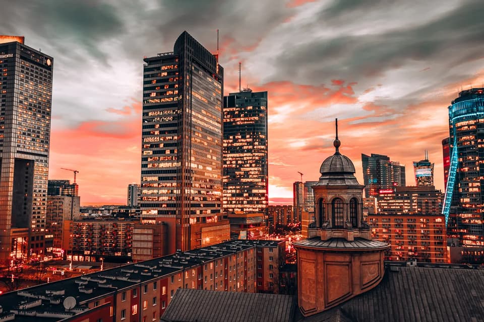Eine moderne Stadtsilhouette mit beleuchteten Wolkenkratzern, die ältere Gebäude überragen, unter einem dramatischen, orange-grauen Sonnenuntergangshimmel.