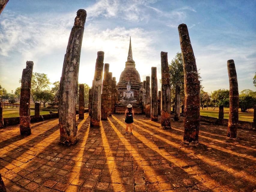 Une personne se tient au milieu de piliers antiques face à une statue de Bouddha et un stupa, avec de longues ombres projetées par le soleil couchant.