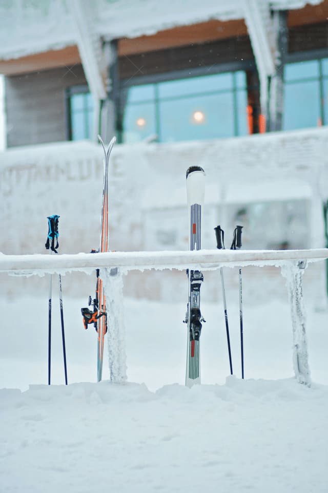 Zwei Paar Skier und Stöcke stehen auf einem eisbedeckten Gestell im Tiefschnee, mit einem verschneiten Gebäude im Hintergrund.