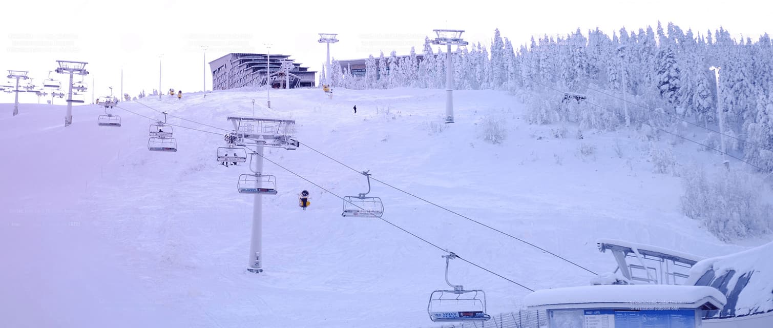 Sessellifte fahren einen schneebedeckten Berg in einem Skigebiet hinauf, mit einem großen Gebäude und einem Wald im Hintergrund.