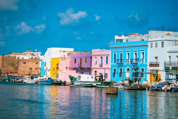 Des bâtiments colorés, jaunes, roses et bleus, bordent un port avec des bateaux amarrés sous un ciel bleu éclatant.