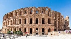 Une vue extérieure d'un grand amphithéâtre romain antique avec trois niveaux d'arches, se dressant sous un ciel bleu clair.